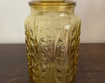 May include: A vintage amber glass jar with a lid. The jar has a decorative embossed pattern of flowers and leaves.