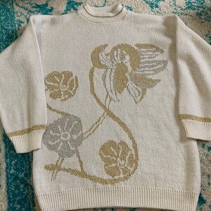 Op de afbeelding: Een witte gebreide trui met een bloemenpatroon in goud- en zilverdraad. De trui heeft een ronde hals en lange mouwen.