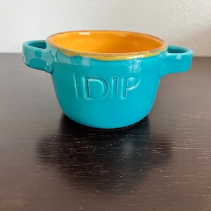 May include: A turquoise ceramic dip bowl with two handles and a yellow interior. The word "DIP" is embossed on the front of the bowl.
