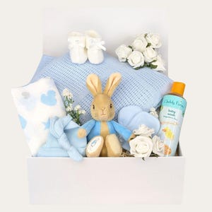 Puede incluir: Una caja de regalo blanca llena de artículos para bebés. Incluye una manta azul, un peluche de Peter Rabbit, patucos, un gorro, un paño, un gel de baño para bebés y rosas blancas. Presentado sobre un fondo blanco.