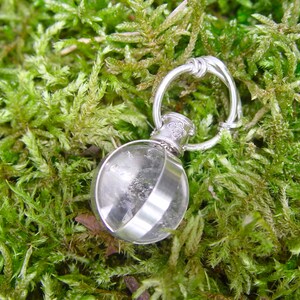 May include: A clear, round glass pendant with a silver-colored metal band and loop. The pendant is filled with a clear substance and rests on a bed of green moss. The loop is knotted at the top, and the top of the pendant has a floral design.