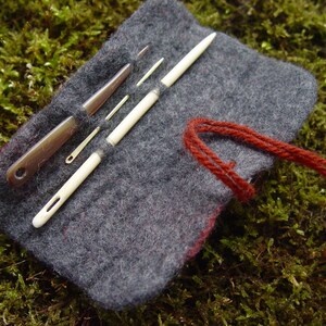 May include: A gray felt needle case with several sewing tools. The case holds a variety of needles and tools, including a brown awl and several white and metal needles. A red thread is attached to the case.