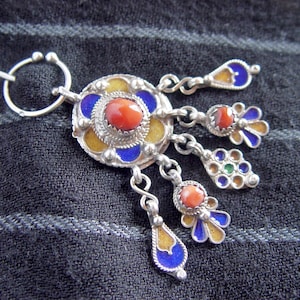 May include: A silver brooch featuring a circular central medallion with a red coral cabochon, surrounded by blue and yellow enamel accents. Four dangling charms hang from the base, each decorated with red coral, blue, yellow, and green enamel.