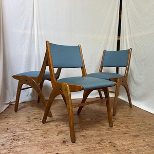 May include: Three mid-century style chairs with wooden frames and blue seats. The chairs feature a distinctive design with angled legs and a curved backrest. The seats are upholstered in a light blue material, and the wooden frames have a warm, natural finish.