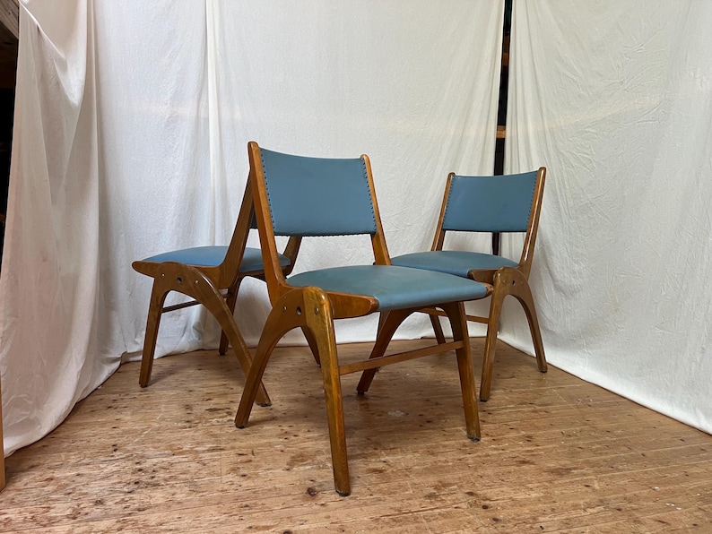 May include: Three mid-century style chairs with wooden frames and blue seats. The chairs feature a distinctive design with angled legs and a curved backrest. The seats are upholstered in a light blue material, and the wooden frames have a warm, natural finish.