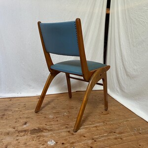 May include: A mid-century modern chair with a blue seat and backrest. The chair has a wooden frame with angled legs and a curved back. The seat and back are upholstered in a light blue material. The chair is set against a white backdrop.