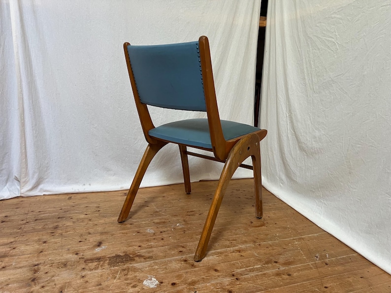 May include: A mid-century modern chair with a blue seat and backrest. The chair has a wooden frame with angled legs and a curved back. The seat and back are upholstered in a light blue material. The chair is set against a white backdrop.