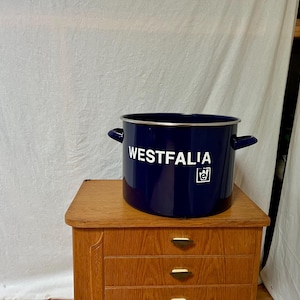 Op de afbeelding: Een marineblauwe emaille pot met een zilveren rand en twee handvatten. Het woord "WESTFALIA" is in het wit op de voorkant van de pot gedrukt, samen met een klein logo. De pot staat op een houten kast.