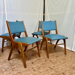 May include: Four mid-century modern chairs with light blue seats and backs. The chairs have wooden frames with a unique angled leg design. The seats and backs are upholstered in a light blue material, and the backs are stitched.