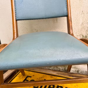 May include: A classic wooden chair featuring a light blue seat and backrest. The chair has a curved design with wooden armrests and legs. The backrest is detailed with small tacks. A vintage piece of furniture.