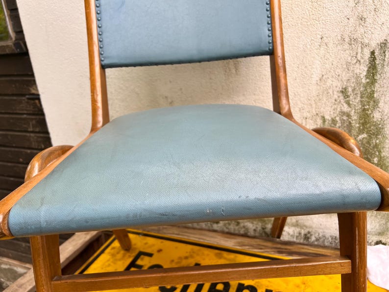 May include: A classic wooden chair featuring a light blue seat and backrest. The chair has a curved design with wooden armrests and legs. The backrest is detailed with small tacks. A vintage piece of furniture.
