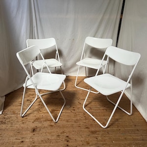 Könnte beinhalten: Vier weiße Klappstühle mit gebogenen Rückenlehnen und Sitzen. Die Stühle haben ein Metallgestell und stehen auf einem Holzboden. Die Stühle sind in einer Gruppe angeordnet, zwei vorne und zwei hinten.