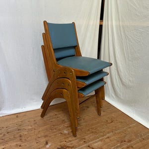 May include: Three wooden chairs stacked together, featuring blue seats and backs. The chairs have a mid-century modern design, with curved wooden frames and a light brown finish. The seats and backs are upholstered in a light blue material.