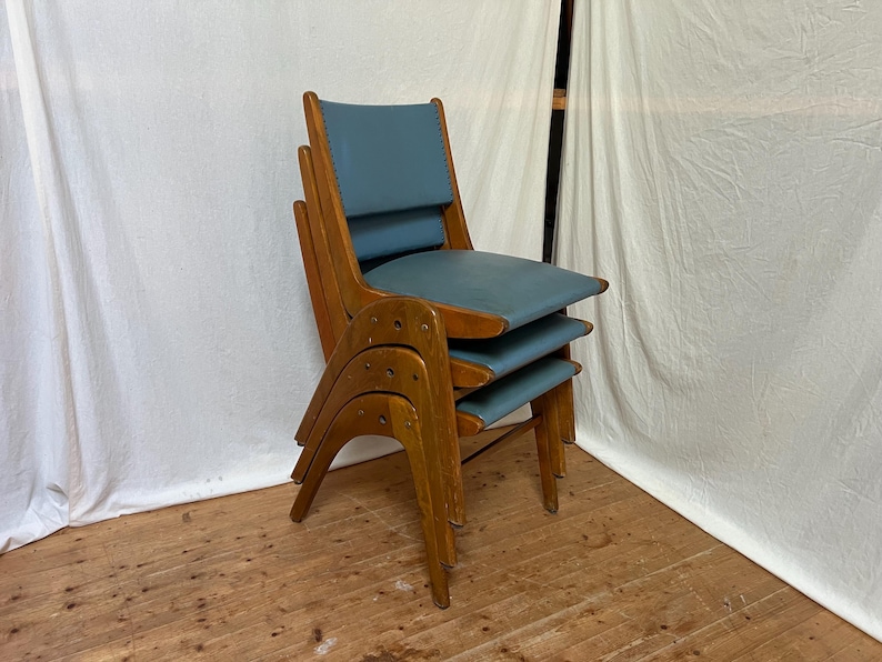 May include: Three wooden chairs stacked together, featuring blue seats and backs. The chairs have a mid-century modern design, with curved wooden frames and a light brown finish. The seats and backs are upholstered in a light blue material.