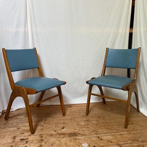 May include: Two mid-century modern chairs with wooden frames and blue leather seats. The chairs have a distinctive curved design and are set against a white backdrop. The seats are approximately 46 cm wide.