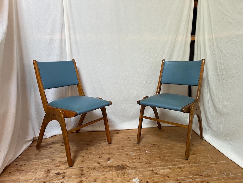 May include: Two mid-century modern chairs with wooden frames and blue leather seats. The chairs have a distinctive curved design and are set against a white backdrop. The seats are approximately 46 cm wide.