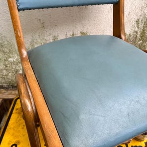May include: Close-up of a vintage chair with a pale blue seat and backrest. The chair has a wooden frame with curved armrests and legs. The seat and backrest are upholstered in a smooth, pale blue material.
