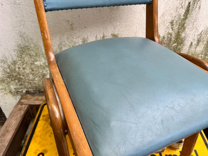 May include: Close-up of a vintage chair with a pale blue seat and backrest. The chair has a wooden frame with curved armrests and legs. The seat and backrest are upholstered in a smooth, pale blue material.