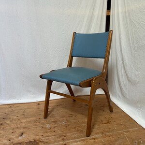 May include: A mid-century modern chair with a light blue seat and backrest. The chair has a wooden frame with a curved back and legs. The seat and back are upholstered in a smooth, solid-coloured material. The chair is set against a white backdrop.