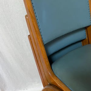 May include: Close-up of a vintage wooden chair with a teal seat and backrest. The chair has a curved back and armrests, with visible wood grain and decorative nailhead trim. The chair's frame is a warm, medium brown colour.