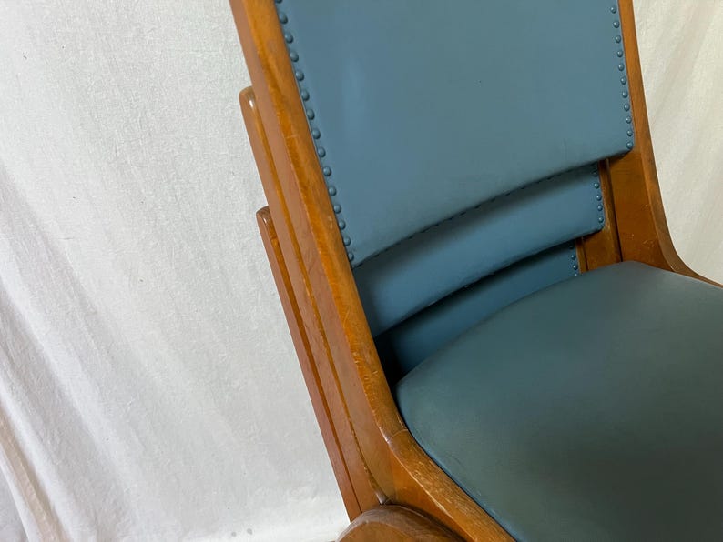 May include: Close-up of a vintage wooden chair with a teal seat and backrest. The chair has a curved back and armrests, with visible wood grain and decorative nailhead trim. The chair's frame is a warm, medium brown colour.