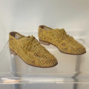 May include: A pair of gold-colored, open-weave lace-up shoes. The shoes have a low heel and a delicate, intricate pattern. They are displayed on a clear, modern table with a white background.