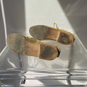 May include: A pair of gold-colored mesh shoes with worn, brown soles. The shoes are displayed on a clear, acrylic surface, showcasing their intricate design and the wear and tear of use. The soles show signs of wear.