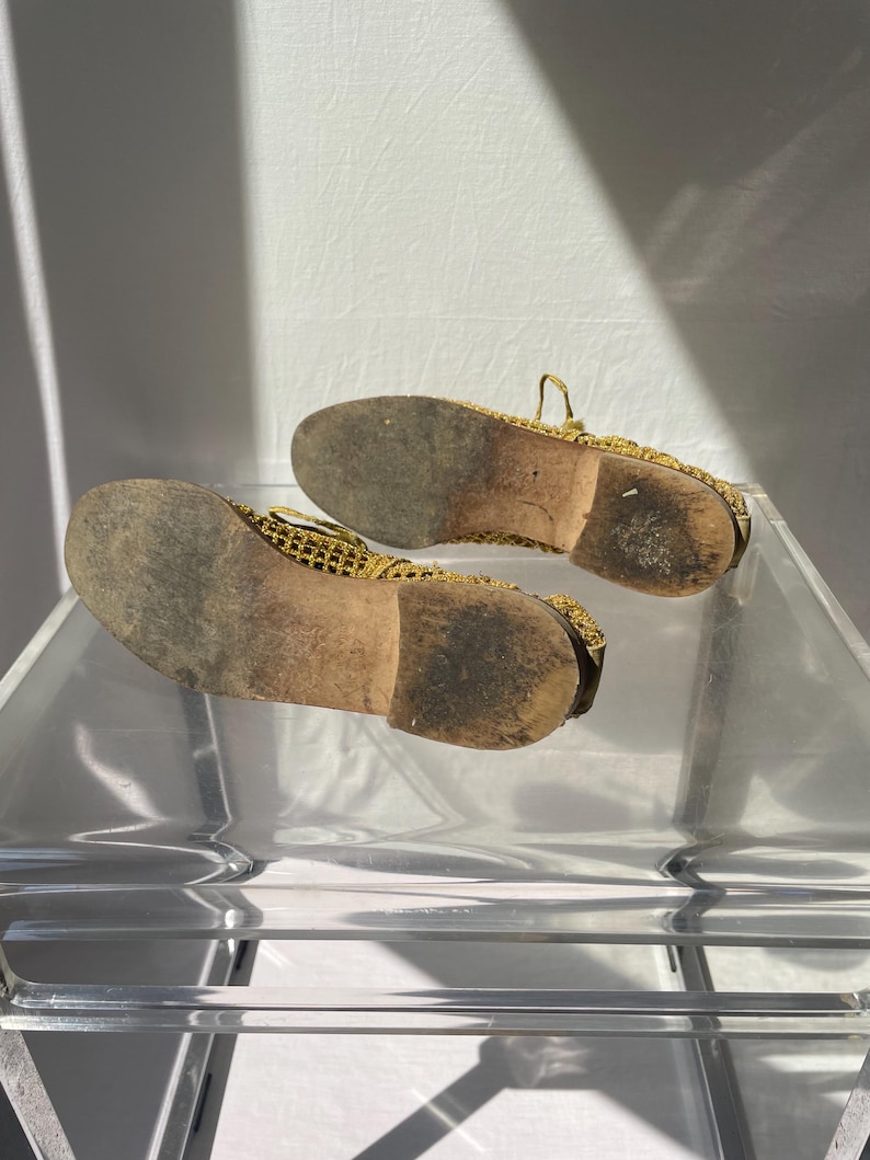 May include: A pair of gold-colored mesh shoes with worn, brown soles. The shoes are displayed on a clear, acrylic surface, showcasing their intricate design and the wear and tear of use. The soles show signs of wear.