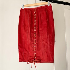 May include: A red pencil skirt with a lace-up front detail. The skirt is made of a soft material and hangs from a wooden hanger. The skirt has a fitted silhouette and a knee-length hemline.