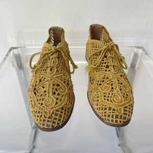 May include: A pair of gold-colored, open-weave crochet shoes with laces. The shoes have a detailed, patterned design and a dark brown sole. The shoes are displayed on a clear surface.