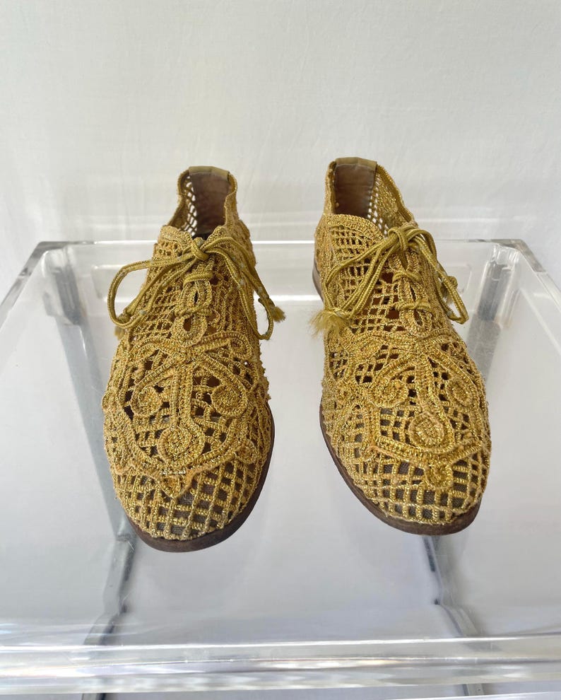 May include: A pair of gold-colored, open-weave crochet shoes with laces. The shoes have a detailed, patterned design and a dark brown sole. The shoes are displayed on a clear surface.