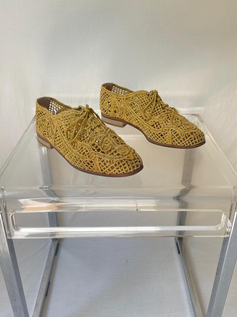 May include: A pair of gold-colored, open-weave lace-up shoes. The shoes have a low heel and a delicate, intricate pattern. They are displayed on a clear, modern table with a white background.