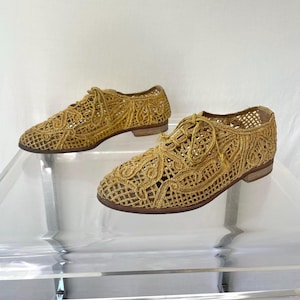 May include: A pair of gold-colored, open-weave shoes with a lace-up design. The shoes have a low heel and a textured, woven appearance. The shoes are displayed on a clear surface.