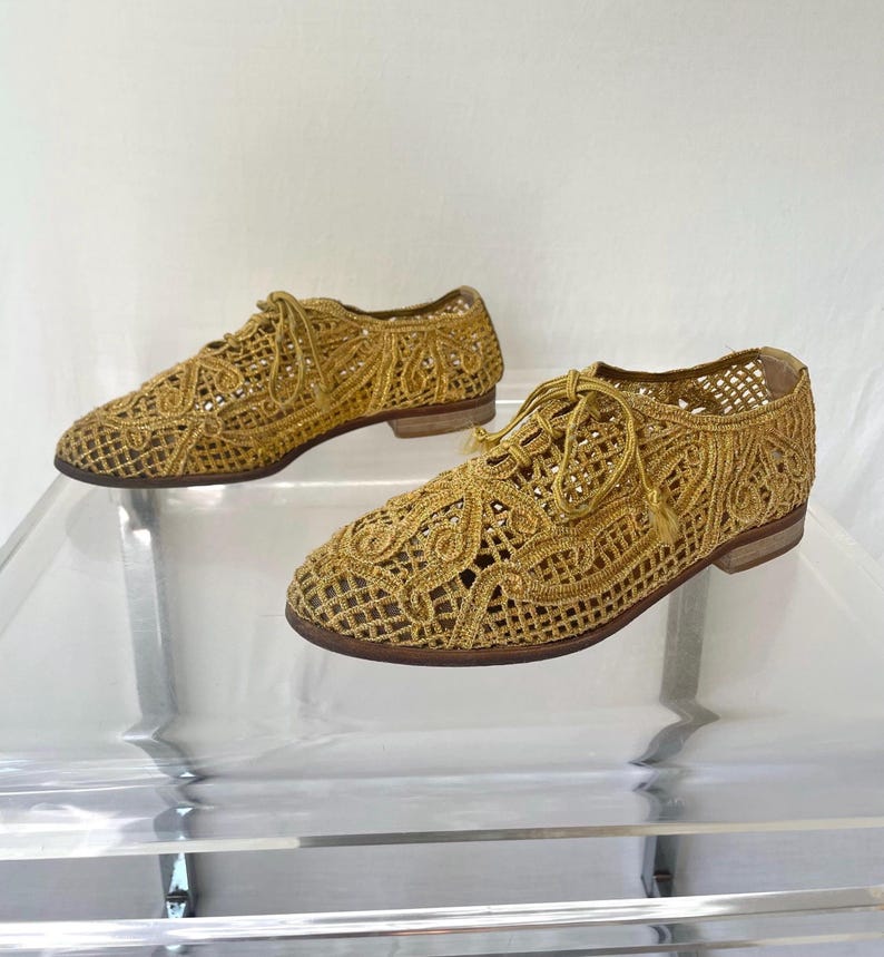 May include: A pair of gold-colored, open-weave shoes with a lace-up design. The shoes have a low heel and a textured, woven appearance. The shoes are displayed on a clear surface.