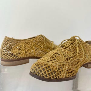 May include: A pair of gold-colored, open-weave shoes with a lace-like design. The shoes have a low heel and a classic Oxford style with laces. The intricate pattern and metallic finish create a unique, elegant look.