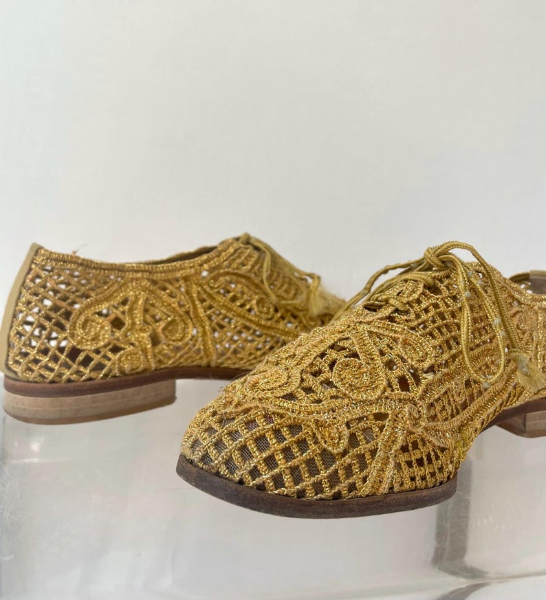 May include: A pair of gold-colored, open-weave shoes with a lace-like design. The shoes have a low heel and a classic Oxford style with laces. The intricate pattern and metallic finish create a unique, elegant look.