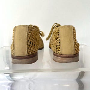 May include: A pair of gold-colored, open-weave shoes with a woven design. The shoes have a rounded toe, a flat sole, and a lace-up closure. The back of the shoe has a solid panel of the same gold color. The shoes are displayed on a clear surface.