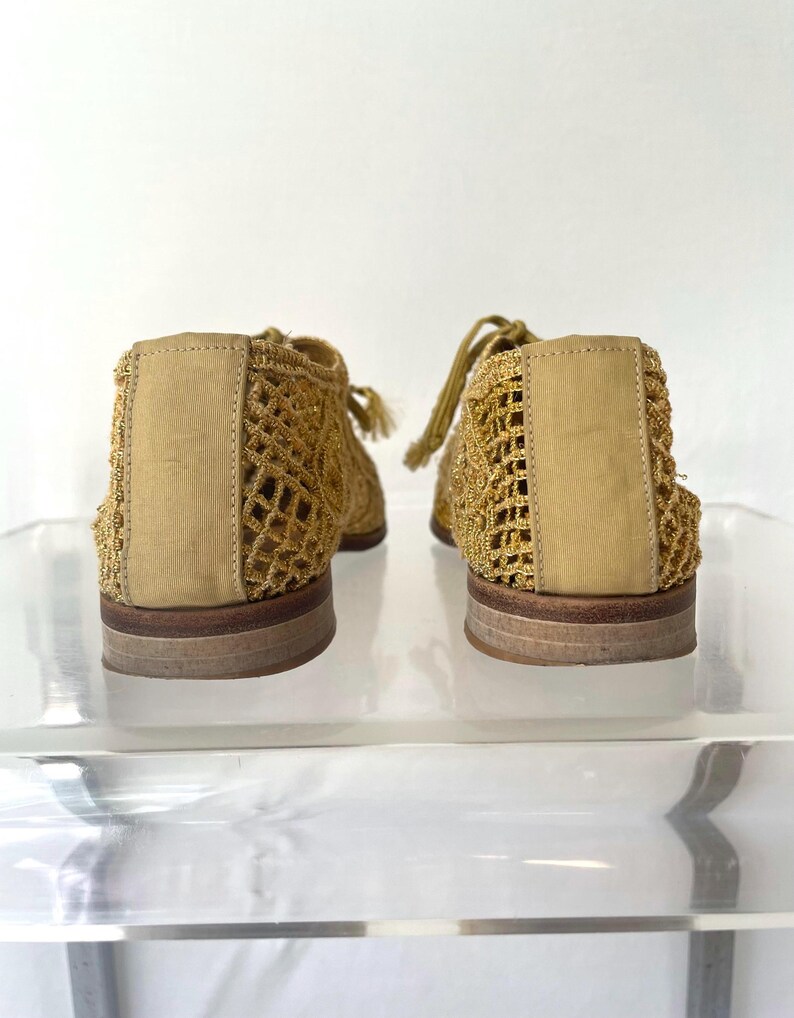 May include: A pair of gold-colored, open-weave shoes with a woven design. The shoes have a rounded toe, a flat sole, and a lace-up closure. The back of the shoe has a solid panel of the same gold color. The shoes are displayed on a clear surface.