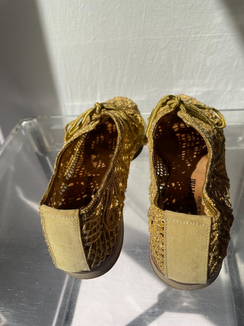 May include: A pair of gold-colored, open-weave shoes with matching laces. The shoes have a rounded toe and a low, flat sole. The material appears to be a metallic thread or yarn, creating a shimmering effect. The shoes are likely for casual wear.