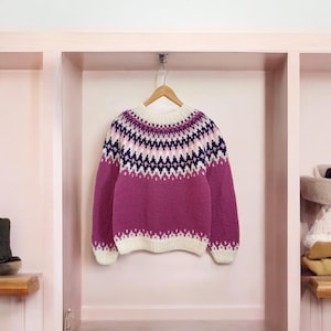 May include: A pink and white knitted jumper with a geometric pattern in navy blue and white. The jumper has a crew neck and long sleeves.