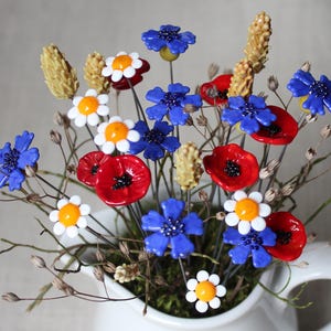 Könnte beinhalten: Ein dekoratives Arrangement aus handgefertigten Glasblumen in einem weißen Keramikkrug. Der Strauß besteht aus leuchtend roten Mohnblumen, blauen Kornblumen und weißen Gänseblümchen mit gelben Mitten, durchsetzt mit strukturierten beigen Weizenähren.