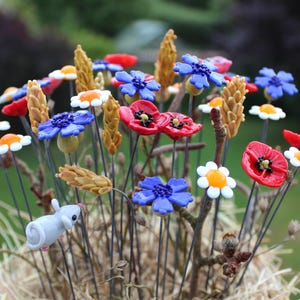 Könnte beinhalten: Eine Ansammlung dekorativer Glasblumenstecker mit roten Mohnblumen, blauen Kornblumen, weißen Gänseblümchen mit orangefarbenen Mitten und goldenen Weizenähren. Eine kleine, hellgraue Glasmausfigur ist an einem der dünnen Metallstiele befestigt.