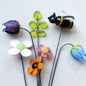 Könnte beinhalten: Eine Sammlung von Gartensteckern aus Glas mit einer violetten Tulpe, einer weißen Blume mit grünem Zentrum, einem grünen Blattstiel, einer kleinen rosa Blume mit gelbem Zentrum, einer orangefarbenen Blume mit schwarzem Zentrum, einer Hummel und einer blauen Glockenblume.