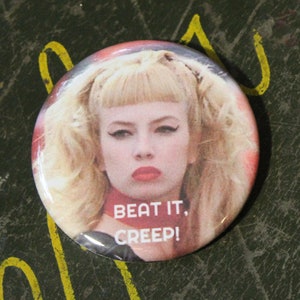 May include: A round button with a woman's face and the text "BEAT IT, CREEP!". The button is next to a quarter coin. The woman has blonde hair and is wearing red lipstick. The background is a dark, textured surface.