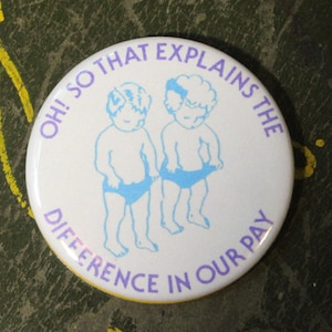May include: A white button with a purple and blue design. Two cartoon figures are depicted with the text "OH! SO THAT EXPLAINS THE DIFFERENCE IN OUR PAY".