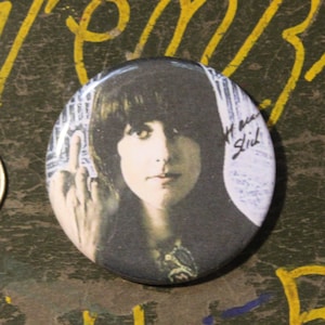 May include: A round button featuring a black and white photo of a woman with long dark hair. The woman is wearing a black shirt and has a serious expression. The button has a white background with a handwritten signature in the upper right corner.