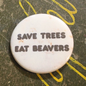 May include: A white button with the text "Save Trees Eat Beavers" in black lettering. The button is on a green surface with yellow markings.
