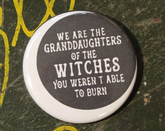 May include: A round pinback button featuring a dark gray circular center with white text that reads: WE ARE THE GRANDDAUGHTERS OF THE WITCHES YOU WEREN'T ABLE TO BURN. A United States quarter coin is placed next to the button for size reference.