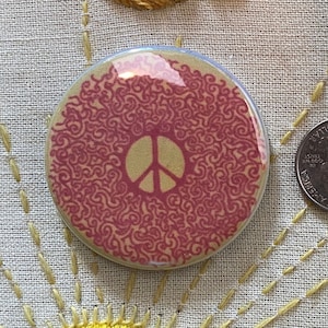 May include: A pink and brown peace sign button with a swirling pattern. The button is about the size of a US quarter.