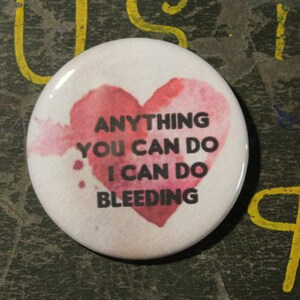May include: A white button with a pink watercolor heart and the text "Anything you can do I can do bleeding".